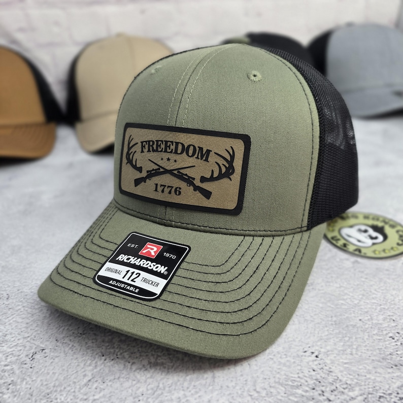 May include: Olive green trucker hat with a black mesh back. The hat features a leather patch with the word "FREEDOM" above "1776" and crossed rifles and antlers. The hat has a curved brim and an adjustable strap.