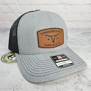 May include: A gray trucker hat with a black mesh back. The front features a brown leather patch with the text "LONGHORN RANCH BLACK ROCK, TX" and a longhorn skull graphic. The hat is adjustable.