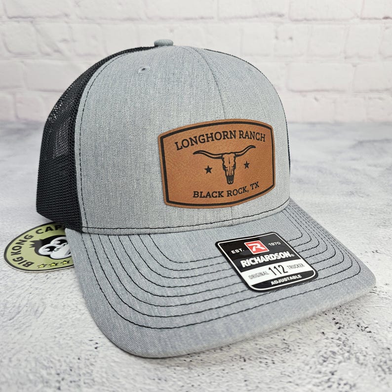 May include: A gray trucker hat with a black mesh back. The front features a brown leather patch with the text "LONGHORN RANCH BLACK ROCK, TX" and a longhorn skull graphic. The hat is adjustable.