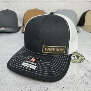 May include: A black Richardson 112 trucker hat with a white mesh back and a curved brim. The hat features the word "FREEDOM" on a rectangular patch. Additional hats in various colors are visible in the background.