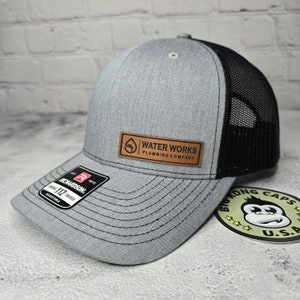 May include: Gray trucker hat with a black mesh back and a curved brim. The hat features a leather patch with the text "WATER WORKS PLUMBING COMPANY". The Richardson brand label is visible on the brim.