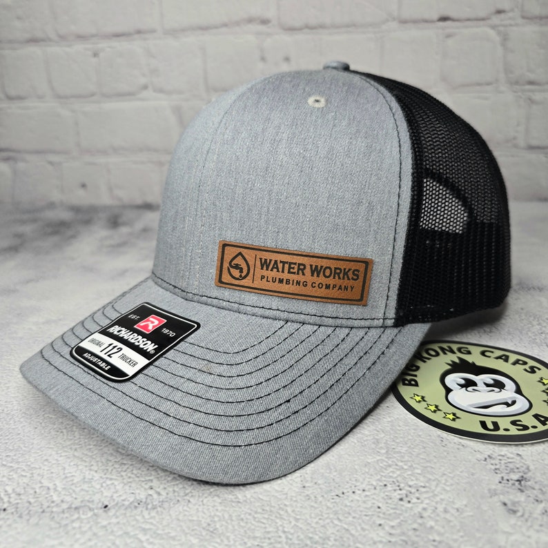 May include: Gray trucker hat with a black mesh back and a curved brim. The hat features a leather patch with the text "WATER WORKS PLUMBING COMPANY". The Richardson brand label is visible on the brim.