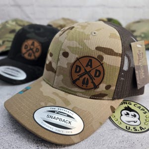 May include: A camouflage print snapback hat with a brown mesh back and a leather patch that says "DAD" with crossed rifles and deer tracks. The hat has a curved brim and a "YUPOONG" tag.