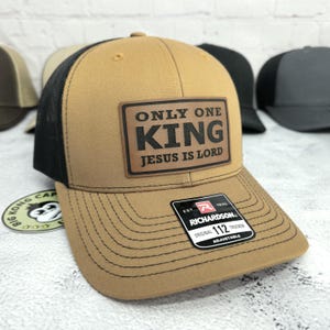 May include: A tan trucker hat with a black mesh back. The hat features a brown leather patch with the text "ONLY ONE KING JESUS IS LORD". The hat has a curved brim and an adjustable strap.