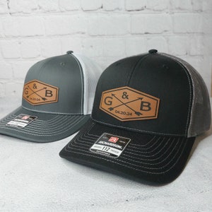 May include: Two black and gray trucker hats with a brown leather patch that says "G & B 04.20.24" with crossed arrows. The hats are both adjustable and have a mesh back.