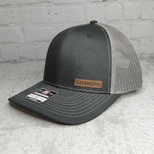 May include: A black and gray trucker hat with a curved brim. The front panel is black, with a brown leather patch that reads "GRANDPA". The back of the hat is gray mesh, and the brim is dark gray with white stitching. The hat has an adjustable strap.