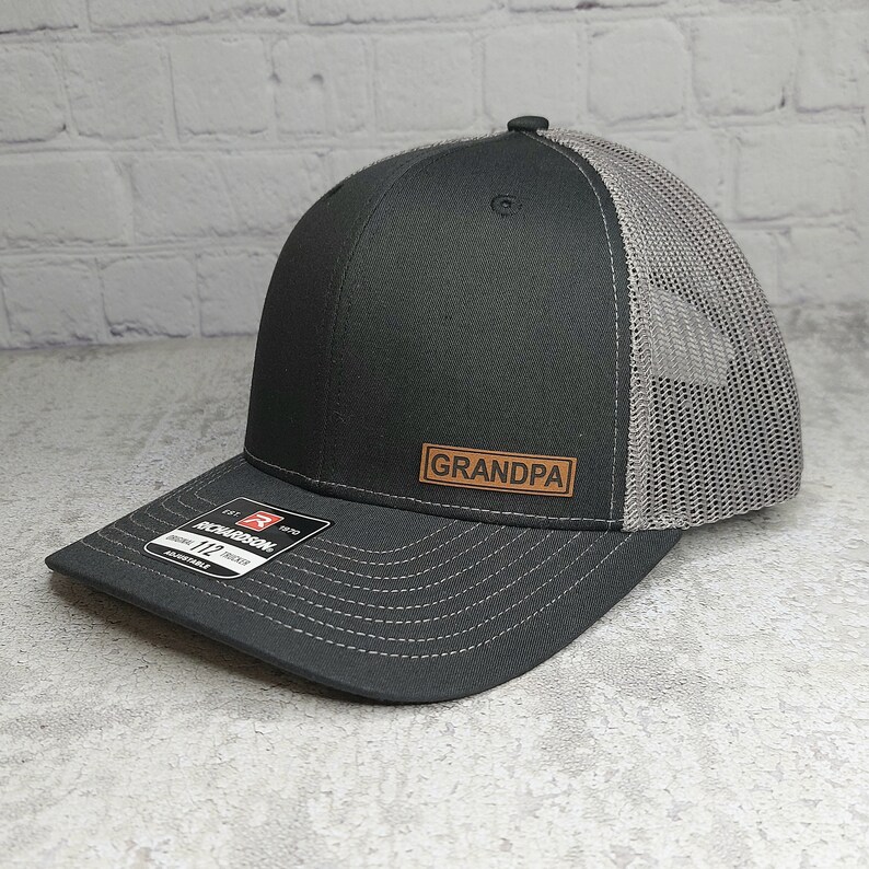 May include: A black and gray trucker hat with a curved brim. The front panel is black, with a brown leather patch that reads "GRANDPA". The back of the hat is gray mesh, and the brim is dark gray with white stitching. The hat has an adjustable strap.