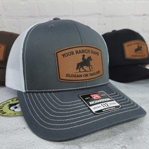 May include: Gray trucker hat with a white mesh back and a brown leather patch. The patch features a silhouette of a rider on a horse and the text "YOUR RANCH NAME SLOGAN OR TAGLINE". The hat has a curved brim and adjustable strap.