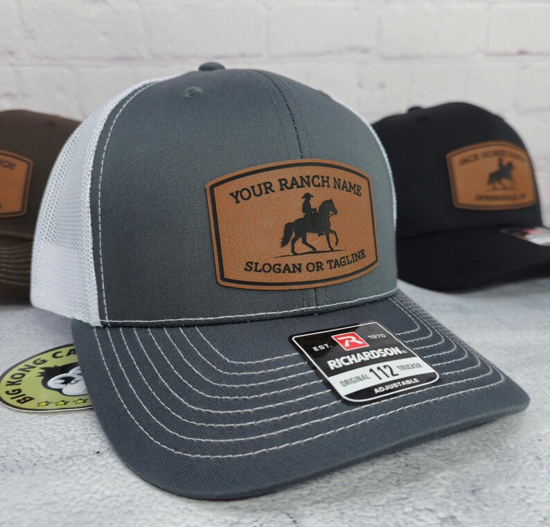 May include: Gray trucker hat with a white mesh back and a brown leather patch. The patch features a silhouette of a rider on a horse and the text "YOUR RANCH NAME SLOGAN OR TAGLINE". The hat has a curved brim and adjustable strap.