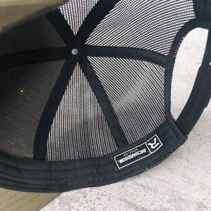 May include: A close-up view of the inside of a trucker hat. The hat features a black mesh back and a black brim. The interior has a black sweatband and a snap closure. The Richardson brand label is visible.