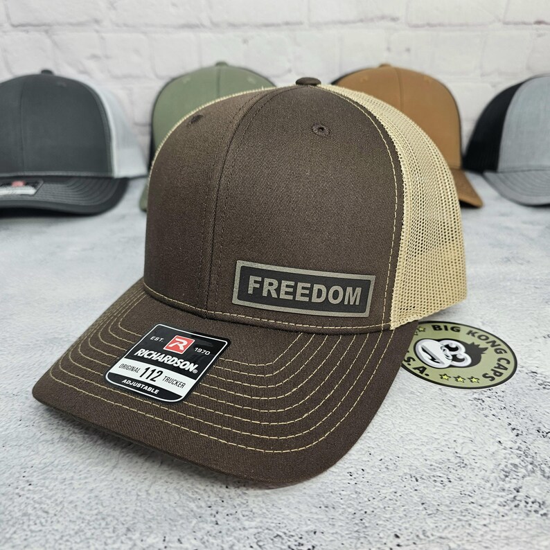 May include: A brown trucker hat with a tan mesh back and a curved brim. The hat features the word "FREEDOM" on a rectangular patch. The hat is adjustable and has a Richardson 112 Trucker tag.