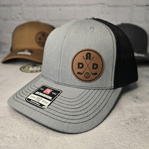 May include: A gray and black trucker hat with a brown leather patch featuring the letters "A D P" and a golf club design. The hat is adjustable and has a Richardson 112 label.