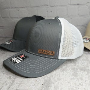 May include: Gray trucker hat with a white mesh back and a brown leather patch that reads "GRANDPA". The hat has a curved brim and an adjustable strap. The Richardson 112 Trucker hat is visible.