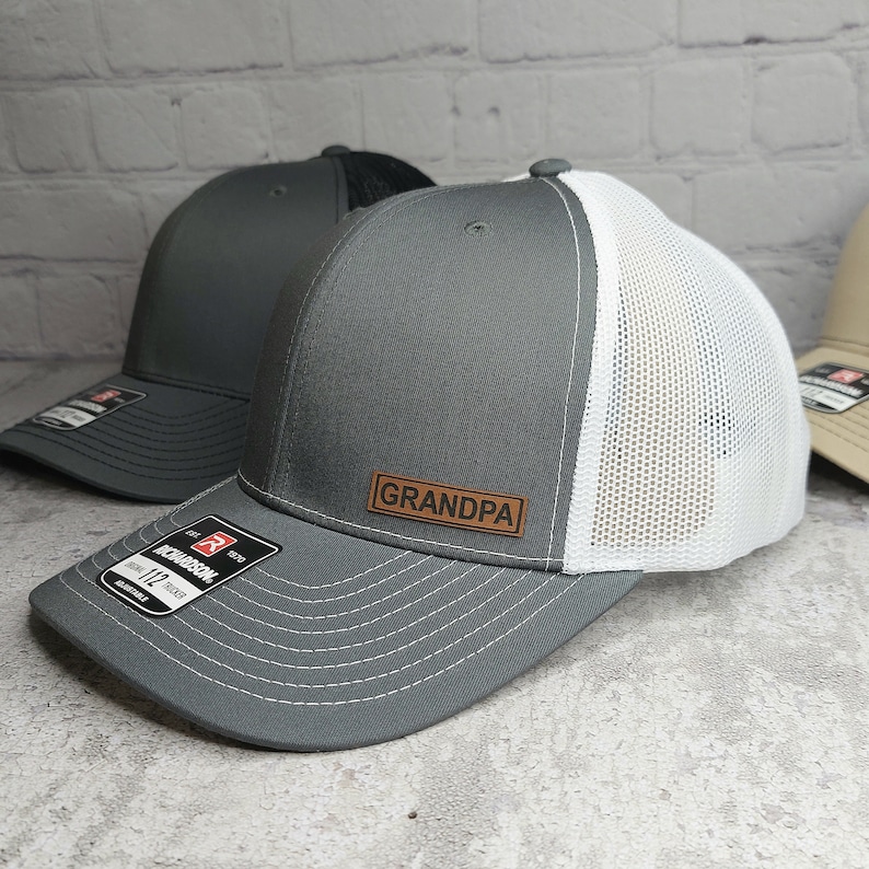 May include: Gray trucker hat with a white mesh back and a brown leather patch that reads "GRANDPA". The hat has a curved brim and an adjustable strap. The Richardson 112 Trucker hat is visible.