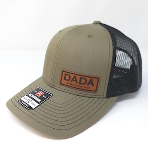 May include: An olive green trucker hat with a black mesh back and a brown rectangular leather patch on the front. The patch features the text DADA and ESTABLISHED 2023 in black. A black sticker on the brim displays the Richardson 112 Trucker logo.