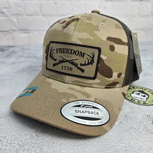May include: A camouflage baseball cap with a black mesh back. The front features a patch with the word "FREEDOM" above crossed rifles and the year "1776". The cap has a curved brim and a snapback closure.