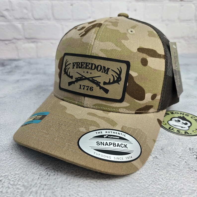 May include: A camouflage baseball cap with a black mesh back. The front features a patch with the word "FREEDOM" above crossed rifles and the year "1776". The cap has a curved brim and a snapback closure.