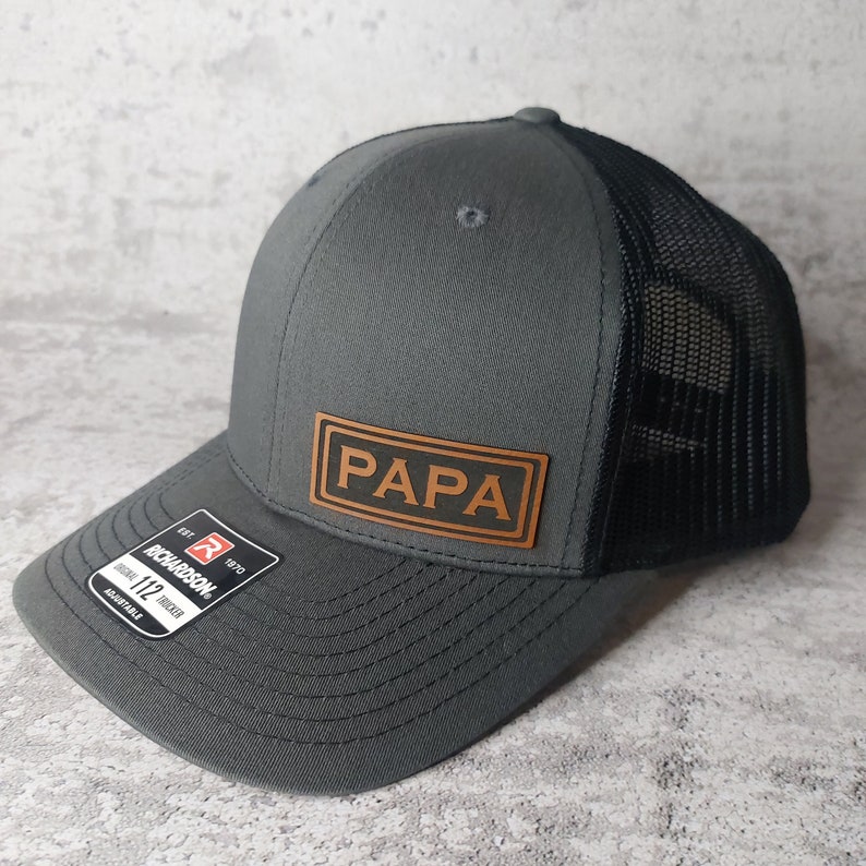 May include: A gray trucker hat with a black mesh back and a brown leather patch that reads "PAPA". The hat has a curved brim and an adjustable strap. The Richardson brand tag is visible on the front.