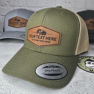 May include: A green and tan trucker hat with a brown leather patch that says "YOUR TEXT HERE CUSTOM TEXT HERE". The patch features a black silhouette of a semi-truck. The hat also has a black and silver sticker that says "THE AUTHENTIC YUPOONG SNAPBACK YUPOONG SINCE 1974".