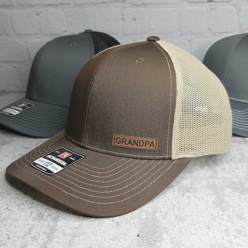 May include: A brown and tan trucker hat with the word "GRANDPA" on a leather patch. The hat has a curved brim and a mesh back. Other hats in gray and black are visible in the background.