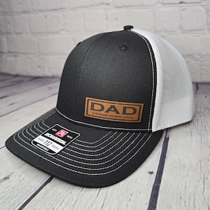 Op de afbeelding: Zwarte en witte truckercap met een bruin leren patch waarop "DAD ESTABLISHED 2024" staat.