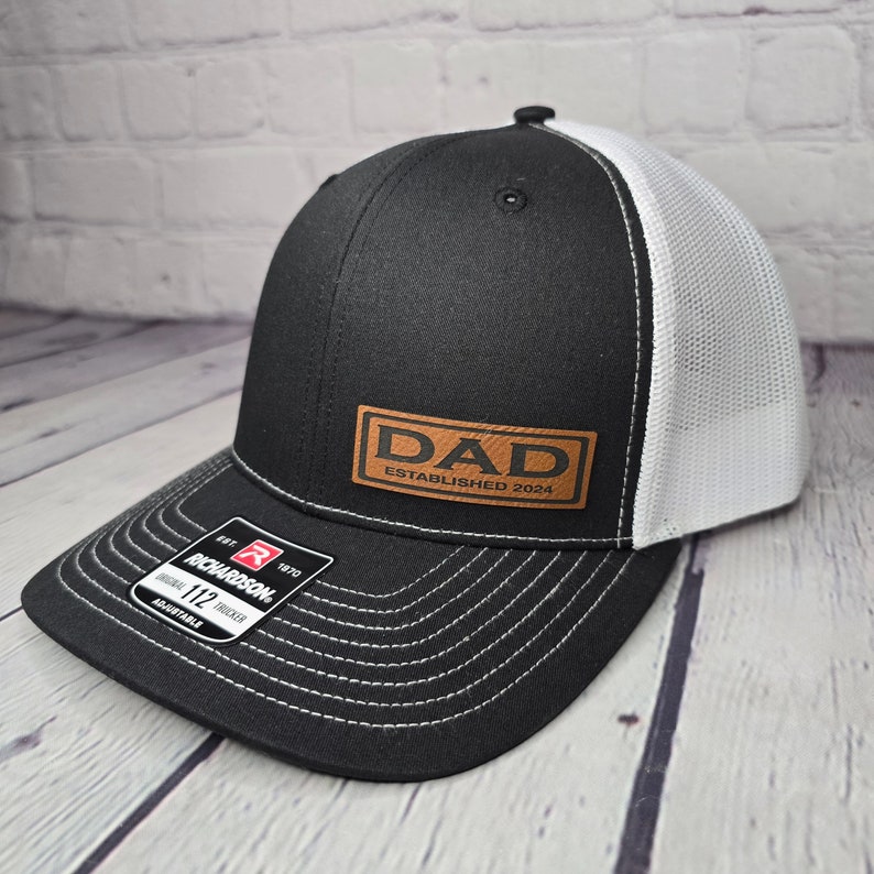 Op de afbeelding: Zwarte en witte truckercap met een bruin leren patch waarop "DAD ESTABLISHED 2024" staat.