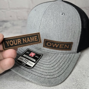 May include: A gray and black trucker hat with a brown leather patch that says "OWEN". The hat also has a small red and white patch that says "EST. 1970 RICHARDSON. ORIGINAL 112 TRUCKER YOUTH".