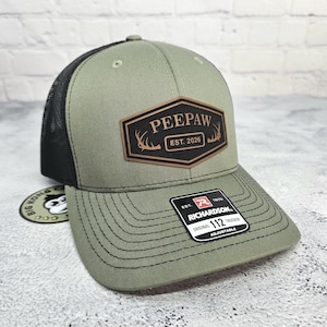 May include: A green and black trucker hat with a brown patch that reads "PEEPAW EST. 2026". The hat has a curved brim and an adjustable strap. The hat also has a Richardson 112 Trucker tag.