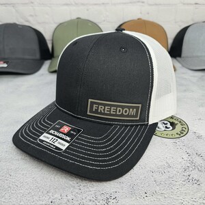 May include: Black and white trucker hat with the word "FREEDOM" on a rectangular patch. The hat has a curved brim with white stitching and a white mesh back. The brand "Richardson" is visible on a tag.
