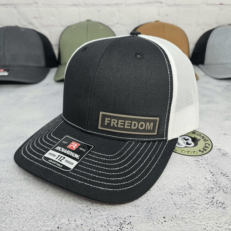 May include: Black and white trucker hat with the word "FREEDOM" on a rectangular patch. The hat has a curved brim with white stitching and a white mesh back. The brand "Richardson" is visible on a tag.