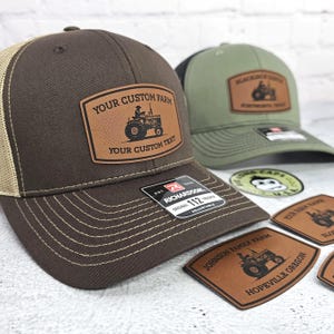 May include: Brown and tan trucker hat with a leather patch featuring a tractor illustration and the text "YOUR CUSTOM FARM" and "YOUR CUSTOM TEXT". A green hat with a black brim is also visible.