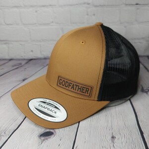 May include: A brown snapback hat with a black mesh back. The hat features the word "GODFATHER" in a black rectangular patch on the front. The brim is curved, and the hat has a Yupoong Snapback sticker.