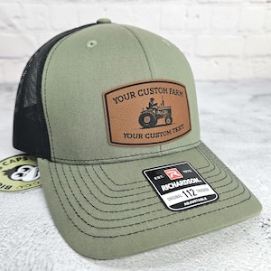 May include: Olive green trucker hat with a black mesh back. The hat features a brown leather patch with a tractor graphic and the text "YOUR CUSTOM FARM" and "YOUR CUSTOM TEXT". The hat is adjustable.