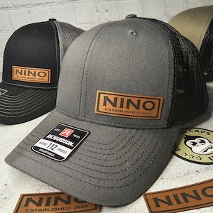 May include: Gray and black trucker hats with a leather patch that reads "NINO ESTABLISHED 2024". The hats have a curved brim and adjustable strap. The Richardson brand label is visible.