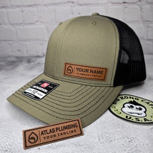 May include: Olive green and black trucker hat with a leather patch that says "YOUR NAME PLUMBING COMPANY". The hat has a curved brim and a mesh back. A sticker with a monkey graphic is also visible.