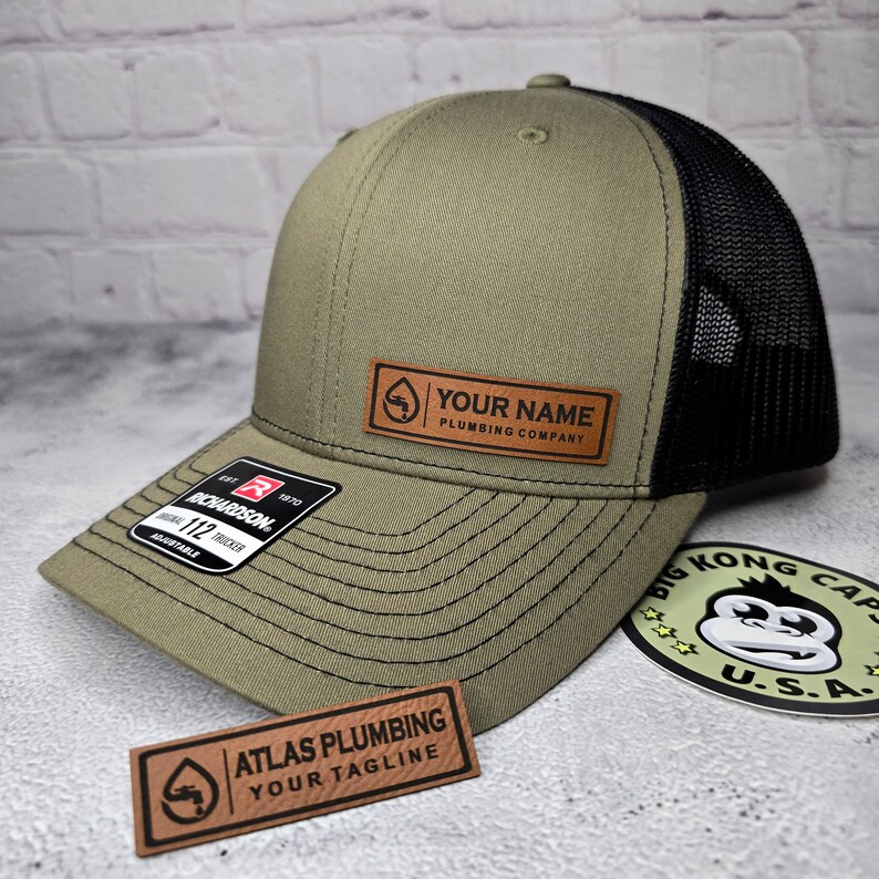 May include: Olive green and black trucker hat with a leather patch that says "YOUR NAME PLUMBING COMPANY". The hat has a curved brim and a mesh back. A sticker with a monkey graphic is also visible.