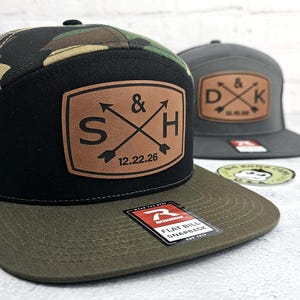 May include: Two flat bill snapback hats. One hat has a camouflage top, black front, and olive green bill. The other is gray. Both have a leather patch with initials and a date. The first hat's patch reads "S & H 12.22.26".