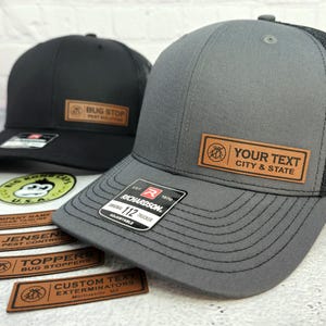 May include: Two trucker hats, one black and one gray, with mesh backs. Each hat has a brown leather patch with custom text. The black hat says "BUG STOP PEST SOLUTIONS." The gray hat says "YOUR TEXT CITY & STATE."