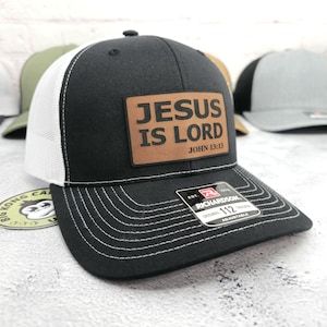 May include: Black trucker hat with a white mesh back and a curved brim. A brown leather patch on the front displays the text "JESUS IS LORD JOHN 13:13". The hat has a Richardson 112 tag and is adjustable.
