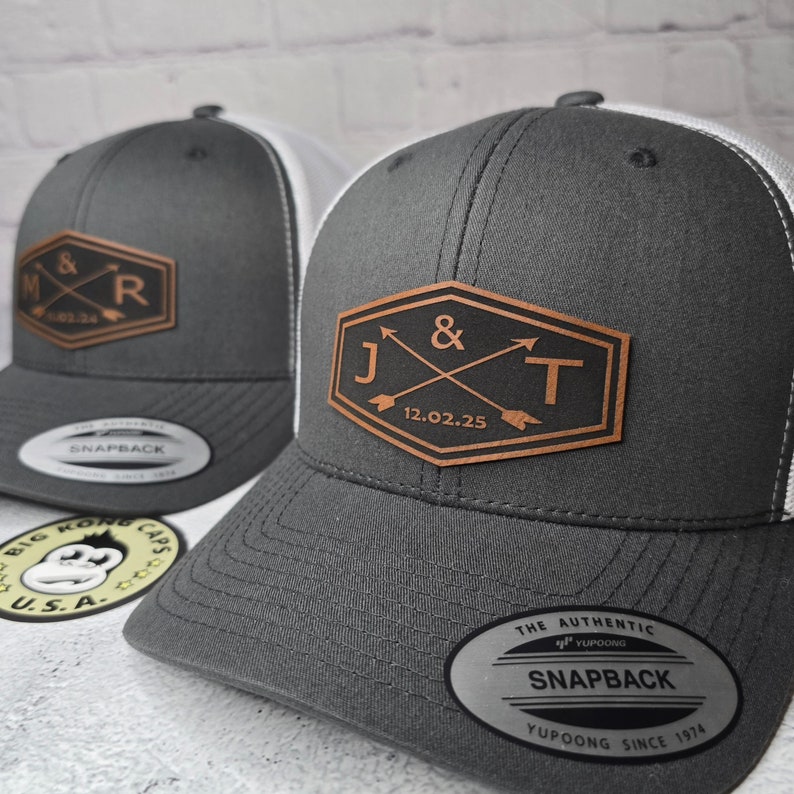 May include: Two gray and white trucker hats with leather patches. The patches feature initials and a date. The first hat has the initials "M & R" and the date "11.02.34". The second hat has the initials "J & T" and the date "12.02.25".