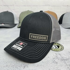May include: Black trucker hat with a gray mesh back and a curved brim. The hat features the word "FREEDOM" on a rectangular patch. The Richardson 112 Trucker hat is adjustable.
