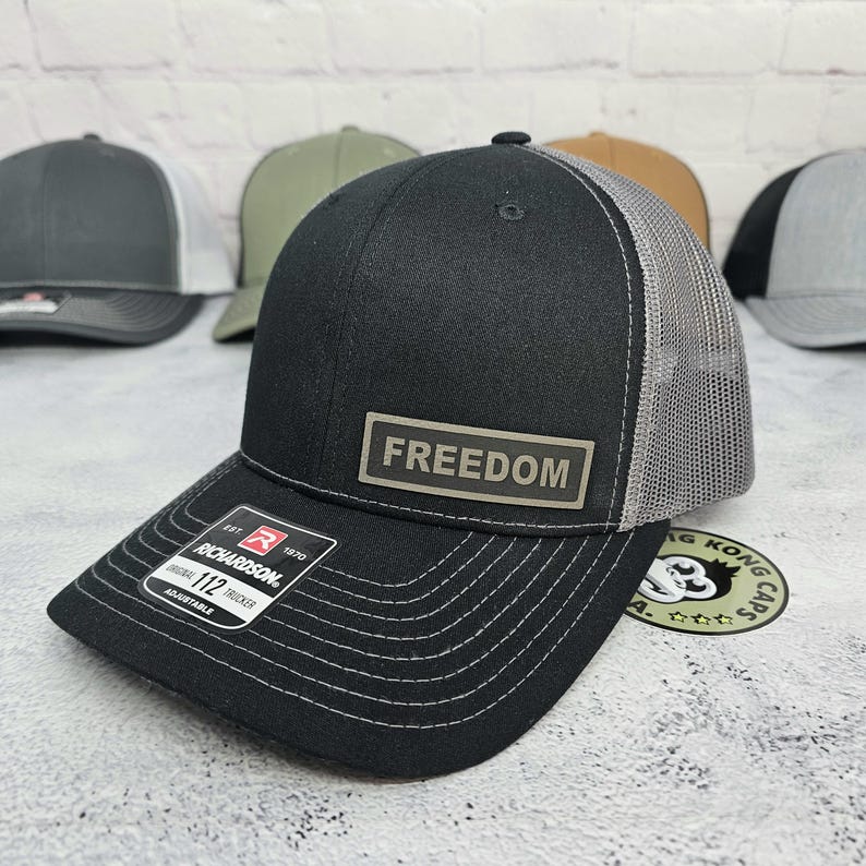 May include: Black trucker hat with a gray mesh back and a curved brim. The hat features the word "FREEDOM" on a rectangular patch. The Richardson 112 Trucker hat is adjustable.