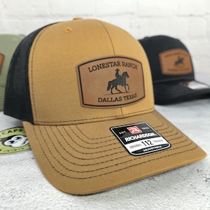 May include: A tan trucker hat with a black mesh back. The hat features a leather patch with the words "Lonestar Ranch Dallas Texas" and a silhouette of a person on a horse. The hat has a curved brim and an adjustable strap.