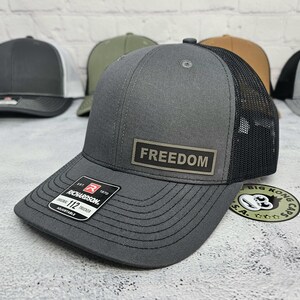 May include: Gray trucker hat with a black mesh back and a curved brim. The hat features a rectangular patch with the word "FREEDOM" and a Richardson 112 tag. Adjustable fit.