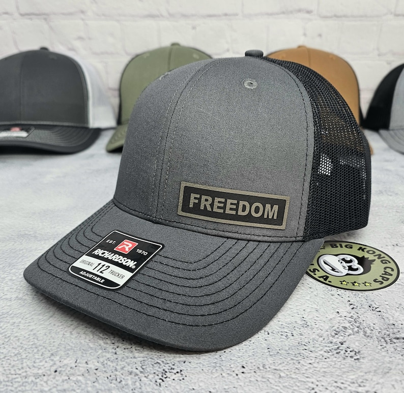 May include: Gray trucker hat with a black mesh back and a curved brim. The hat features a rectangular patch with the word "FREEDOM" and a Richardson 112 tag. Adjustable fit.