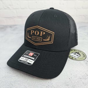 May include: Black trucker hat with a mesh back and a curved brim. The front features a brown hexagon patch with the word "POP" and "EST. 2026" printed on it. The hat is adjustable and has a Richardson tag.