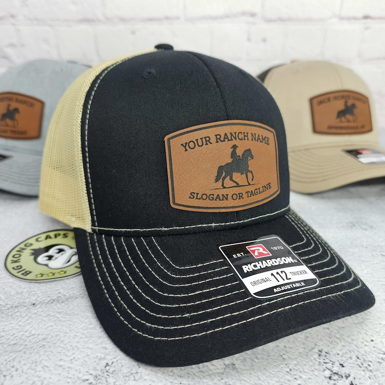 May include: Black trucker hat with a tan mesh back and a leather patch. The patch features a silhouette of a rider on a horse and the text "YOUR RANCH NAME" and "SLOGAN OR TAGLINE". The hat is adjustable.