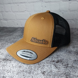 May include: A tan and black trucker hat with the word "Abuelo" in brown lettering. The hat has a curved brim and a mesh back. The hat has a snapback closure and a Yupoong label.