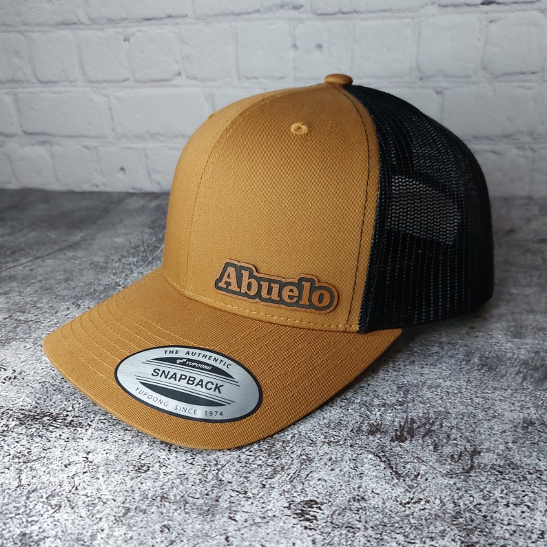 May include: A tan and black trucker hat with the word "Abuelo" in brown lettering. The hat has a curved brim and a mesh back. The hat has a snapback closure and a Yupoong label.