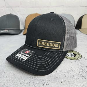 May include: Black trucker hat with a gray mesh back and a curved brim. The hat features a rectangular patch with the word "FREEDOM" in gold lettering. The hat has a Richardson 112 Trucker adjustable tag.